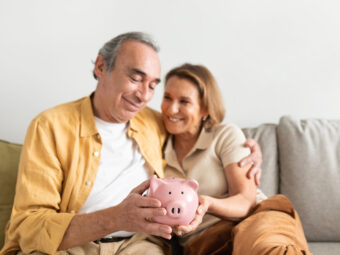 Happy european senior couple holding piggybank and embracing, sitting on sofa in living room interior, free space